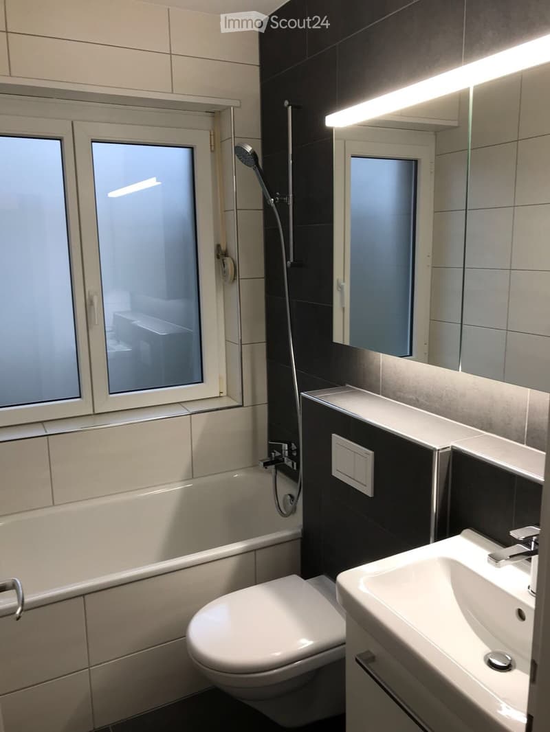 Modern bathroom with a toilet, sink, mirror, and showerhead. The walls are tiled in a combination of light beige and dark black tiles, and there is a window with frosted glass above the bathtub.