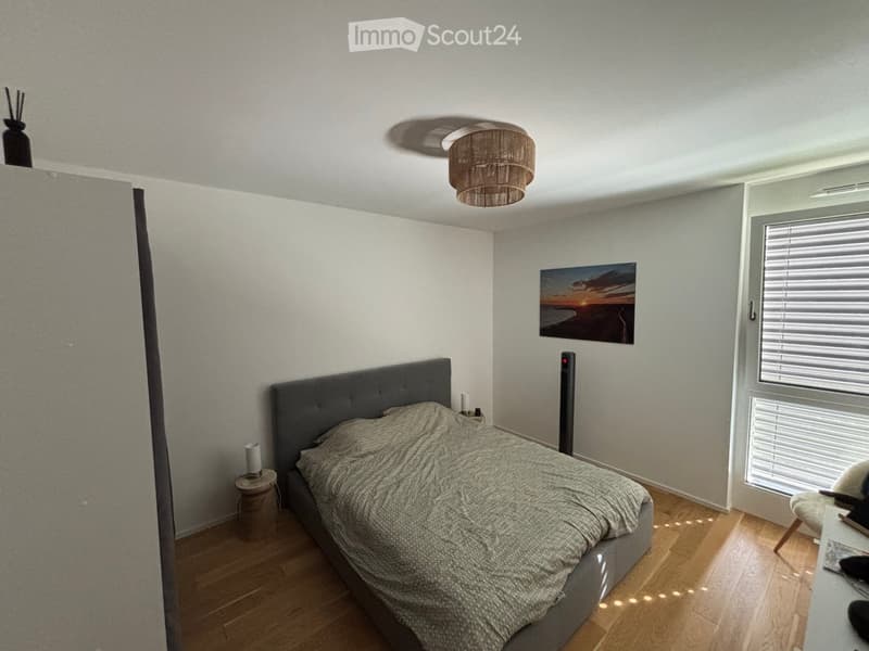 Modern bedroom with a grey upholstered bed, wooden floor, nightstand, table lamp, picture frame, and a ceiling light fixture.