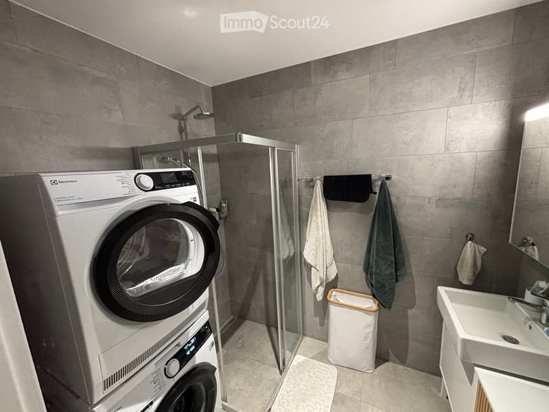 Modern bathroom with a washing machine, sink, mirror, shower, and towels.