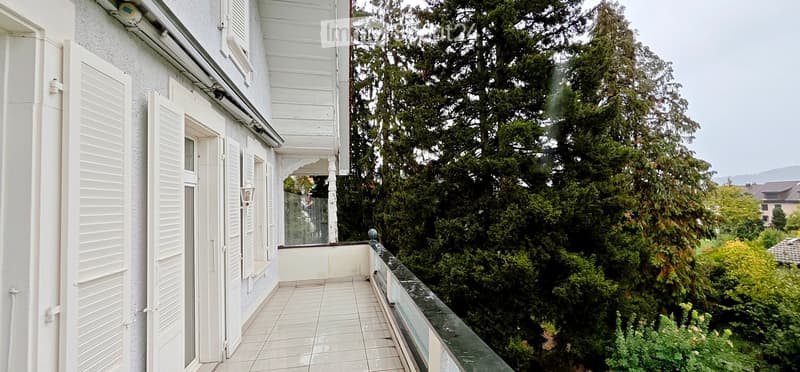 Balcony with white tiled flooring, glass railing, and view of lush green trees and distant hills.