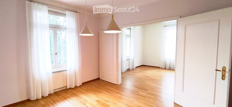 Empty room with wooden flooring, white walls, large windows with white curtains, and pendant lights.