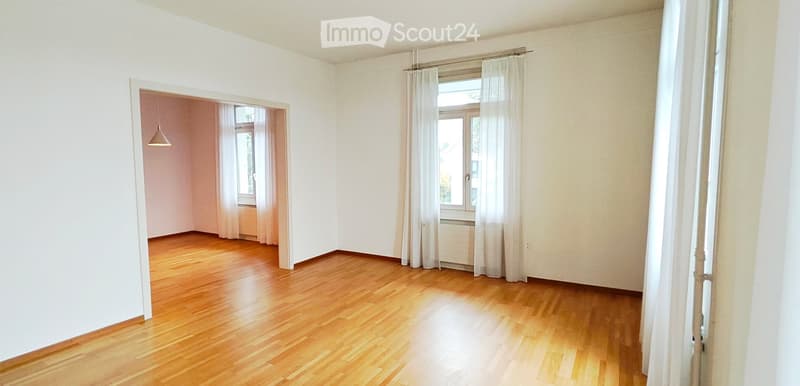 Empty room with wooden flooring, white walls, and large windows with white curtains. A doorway leads to another room with similar features.