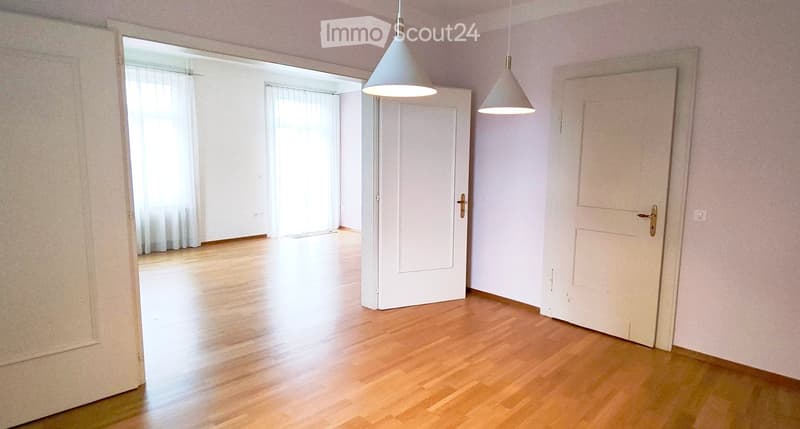 Empty room with light wooden flooring, white walls, and two pendant lights. There are two doors and a window with white curtains visible in the background.