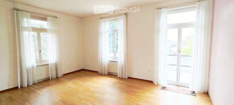 Empty room with wooden flooring, white walls, and large windows with white curtains. The room has a sliding glass door leading to a balcony.