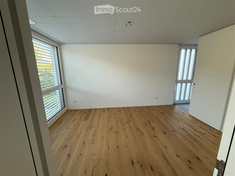 Empty room with wooden flooring, white walls, large windows with blinds, and a door leading to another space.