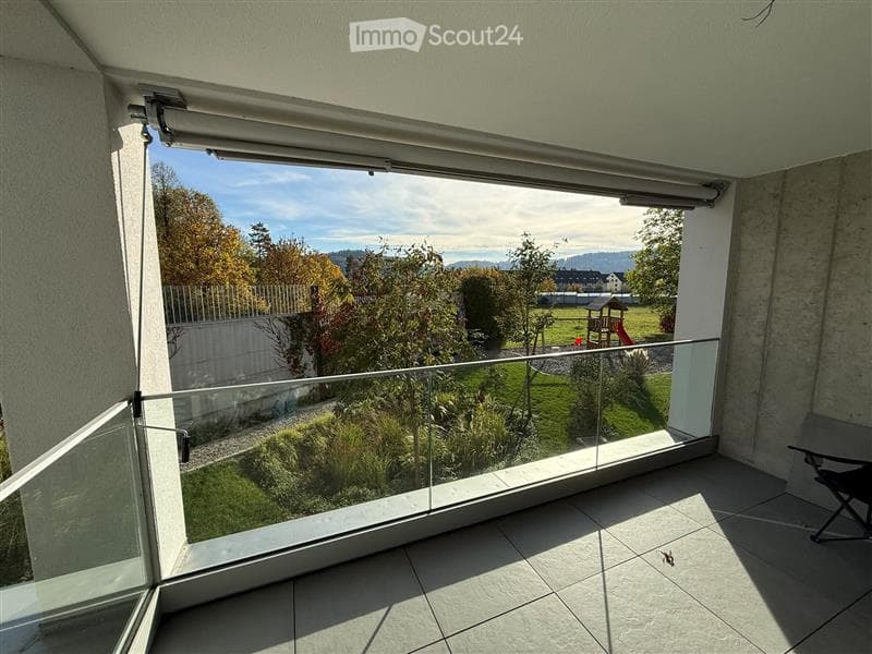 Balcony with large glass sliding doors, tiled floor, and a view of a landscaped garden with trees and a playground.