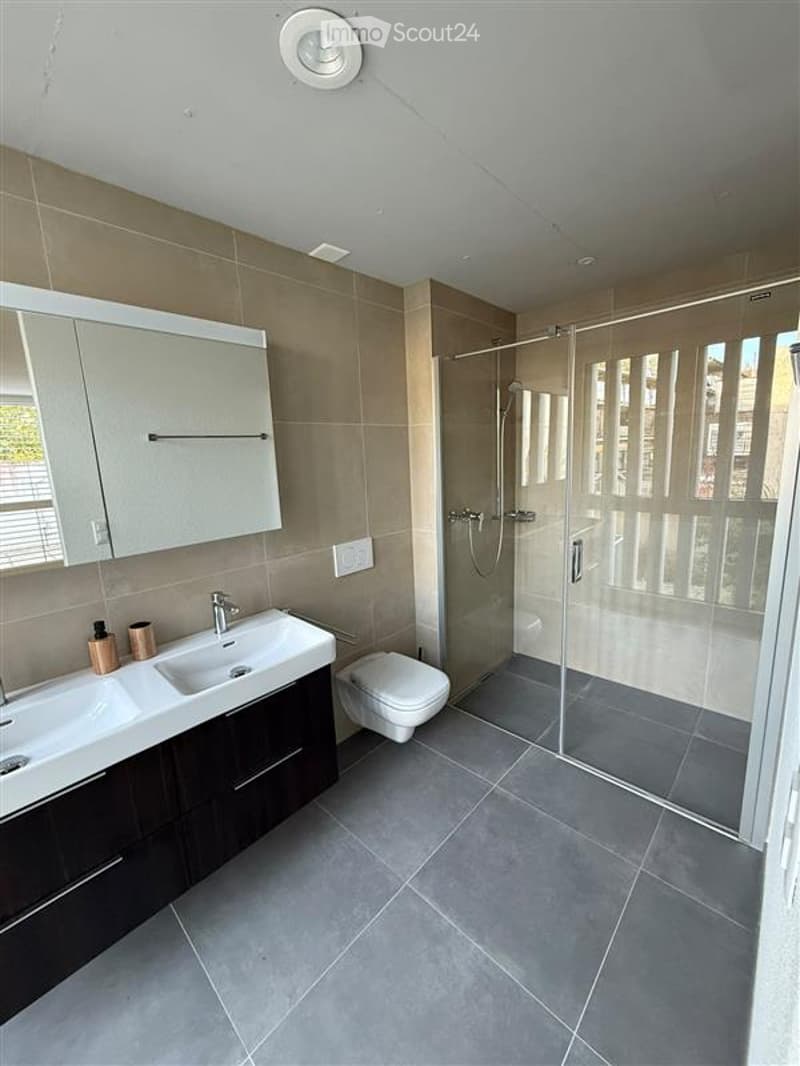 Modern bathroom with double sink, large mirror, toilet, and glass-enclosed shower. Features dark wood cabinetry and grey tiles.