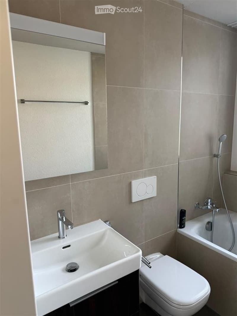 Modern bathroom with a white sink, mirror, toilet, bathtub, and showerhead. Beige tiled walls and a sleek design.