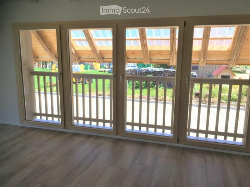 Empty room with wooden flooring and large sliding glass doors leading to a covered outdoor area with wooden railings and a view of a green lawn.