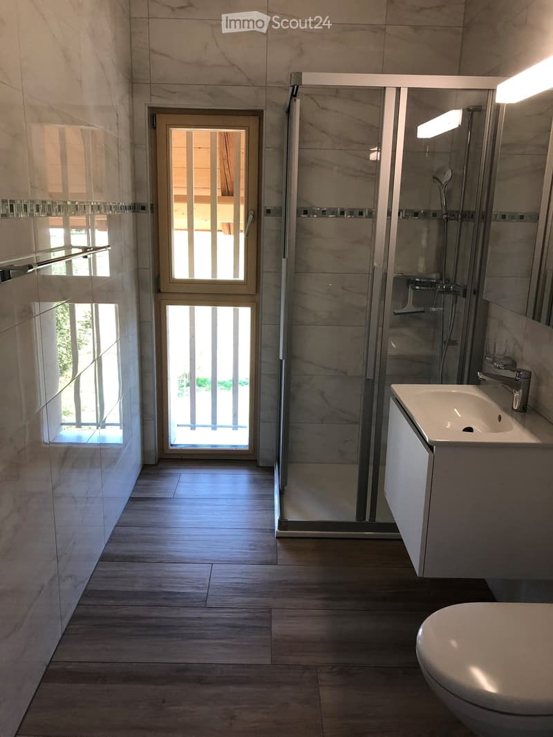 Modern bathroom with marble tiles, a glass-enclosed shower, a sink with a modern faucet, and a toilet. The room features large windows and a door leading outside.