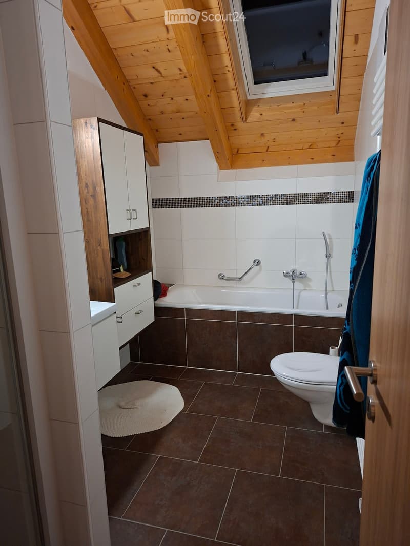 Bathroom with a bathtub, toilet, white cabinets, and a towel hanging on the door. The room features brown tiled flooring and a skylight window.