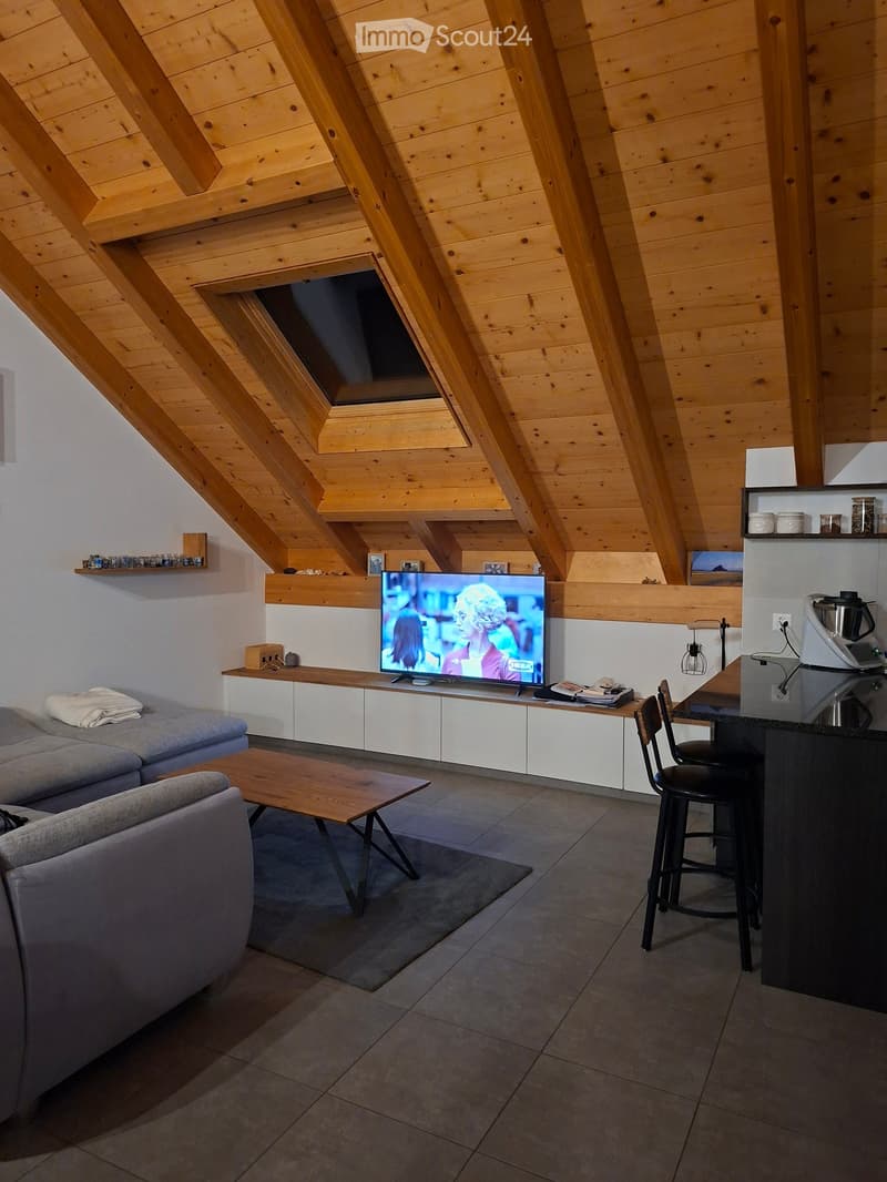 Modern living room with exposed wooden beams, grey sofa, coffee table, television, and kitchen counter with stools.