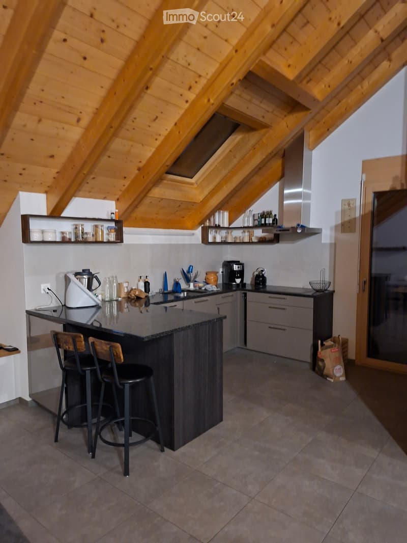 Modern kitchen with dark wooden island, two bar stools, stainless steel appliances, and open shelving displaying jars and bottles.
