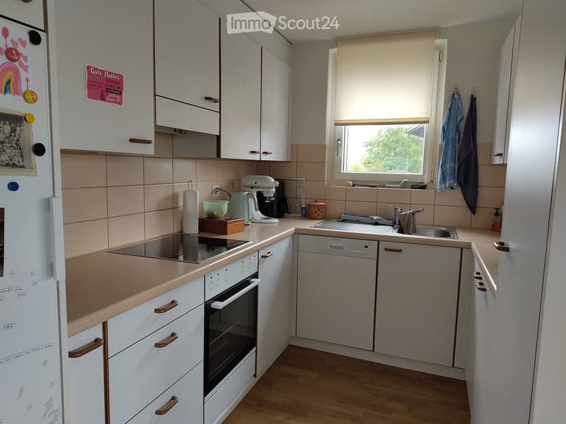 Modern kitchen with white cabinets, light wooden countertops, and various appliances including a refrigerator, oven, dishwasher, and mixer. The space features a window with blinds and a tiled backsplash.