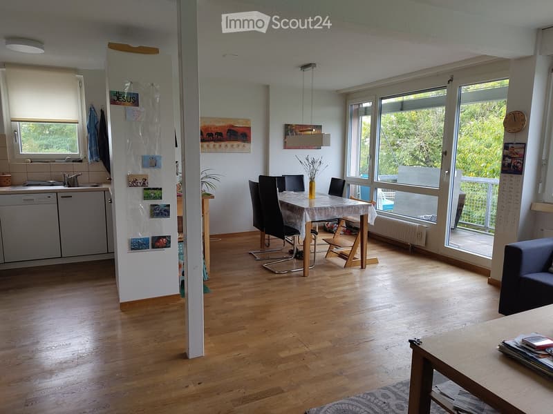 Open-plan living and dining area with wooden flooring, large windows, a sofa, coffee table, dining table with chairs, and a kitchen area with a dishwasher and sink.