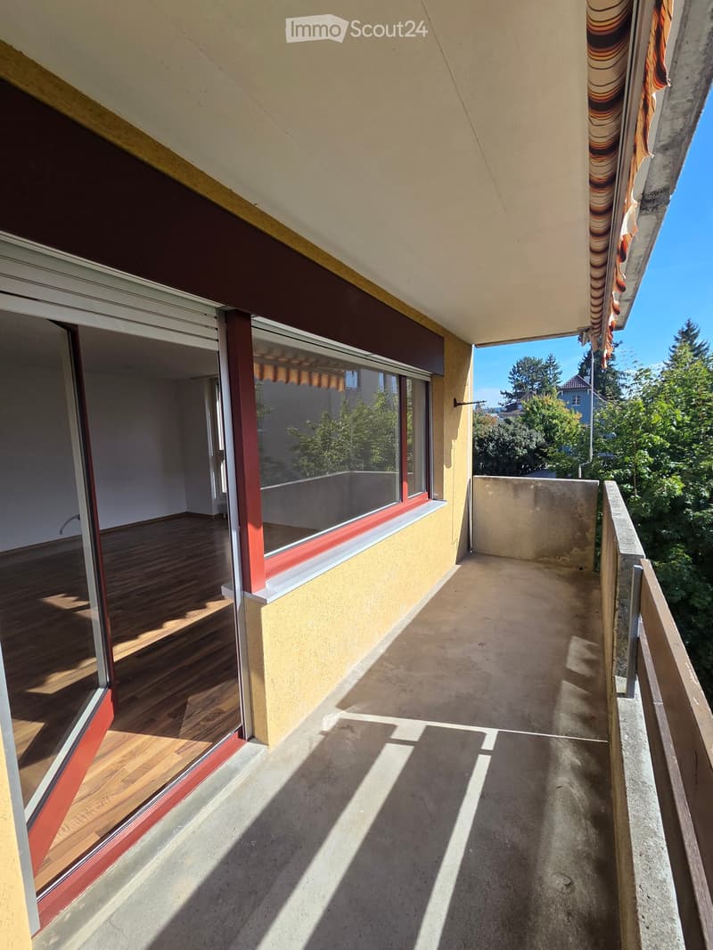 Balcony with wooden flooring, large windows, and a view of greenery and trees.
