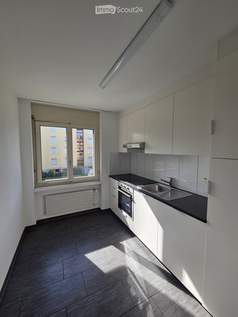 Modern kitchen with white cabinets, dark countertops, stainless steel sink, and built-in oven. Features a large window with a view of the building exterior.