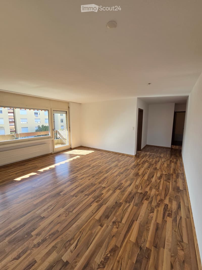 Empty room with wooden flooring, white walls, and large windows with a view of the outside. The room has a door leading to another area and a radiator under one window.