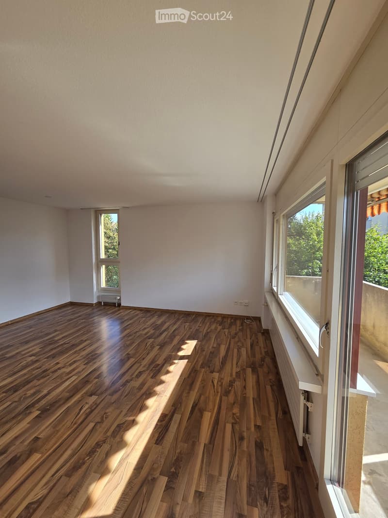 Empty room with wooden flooring, white walls, and large windows allowing natural light to enter.