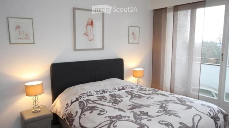 Modern bedroom with a large bed, nightstands, table lamps, and artwork on the walls. Large windows with sheer curtains allow natural light to enter.