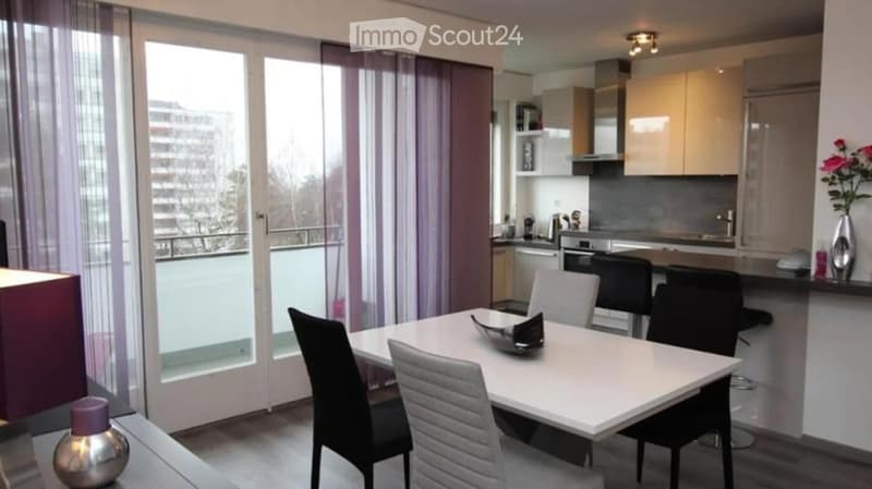 Modern, open-plan living and dining area with a white dining table, chairs, and a kitchen area featuring cabinets, an oven, and a dishwasher. Large windows with sheer curtains offer a view of the balcony.