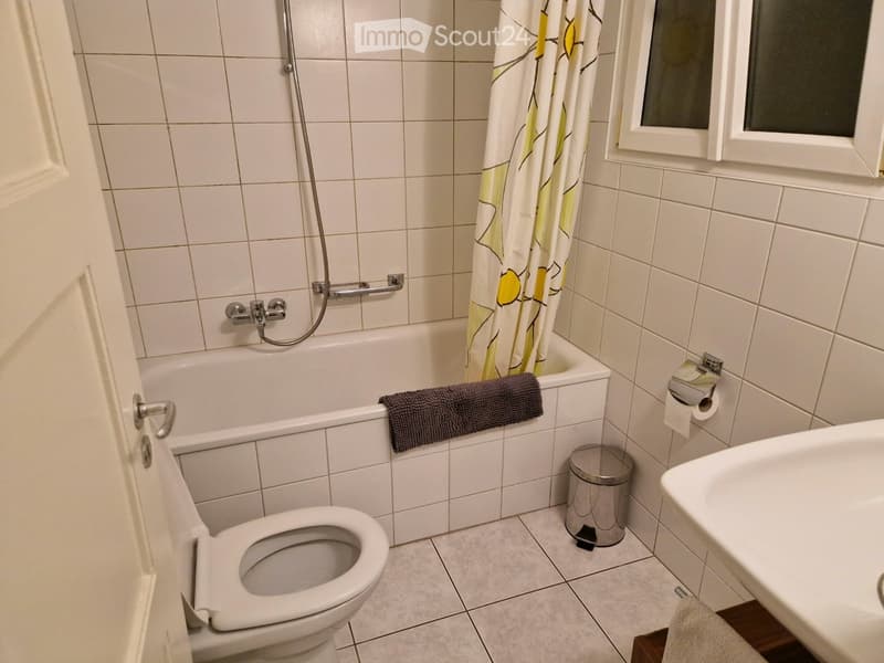 Bathroom with white tiled walls and floor, featuring a bathtub with a shower curtain, a toilet, a sink, a towel, and a trash can.