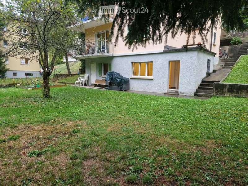 Lush green lawn with a small tree, a covered outdoor seating area with chairs and a table, and a wooden bench. A modern house with a balcony and stairs leading up to it is visible in the background.