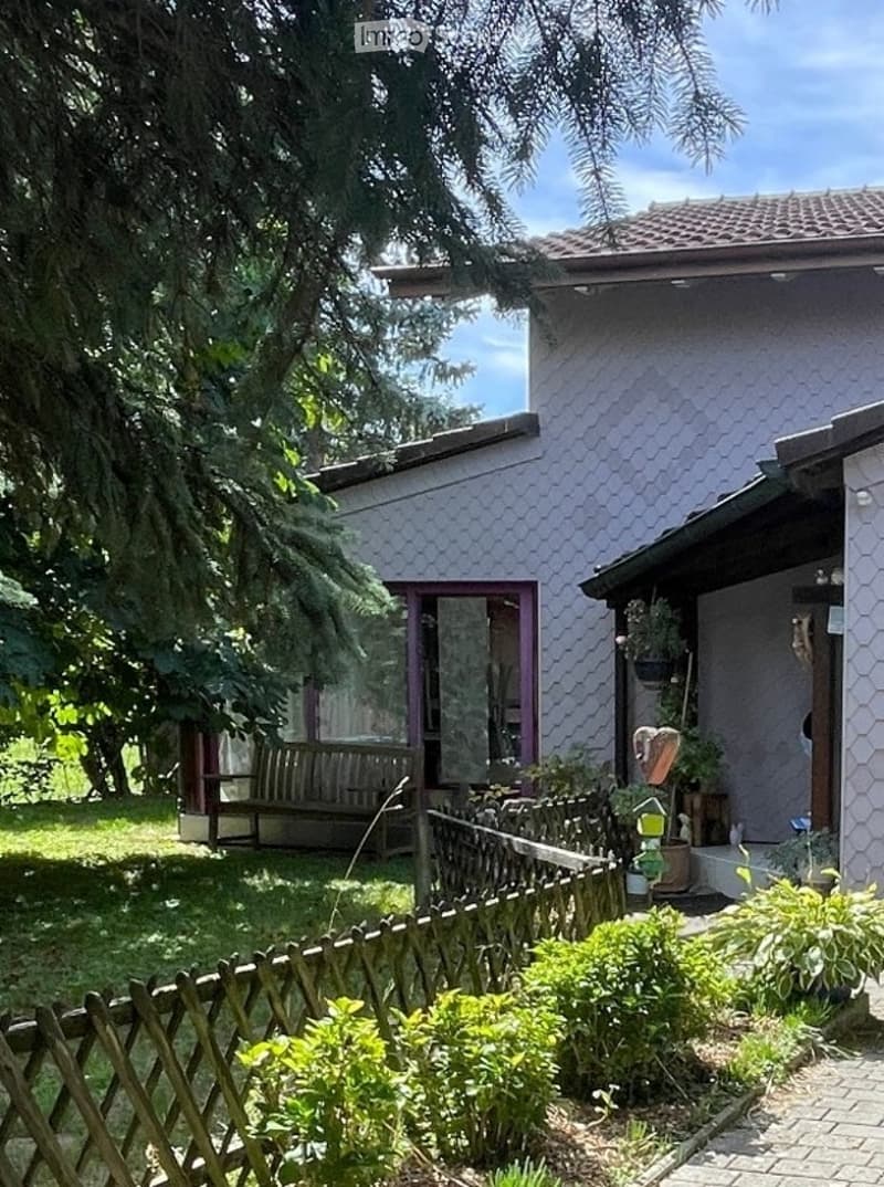 A peaceful garden area with a wooden fence, a bench, and various plants and trees surrounding a house with a tiled roof and purple accents.