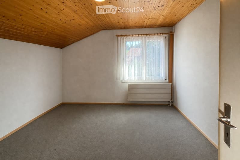 Empty room with light-colored walls, carpeted floor, wooden ceiling, window with white curtains, and a radiator below the window.