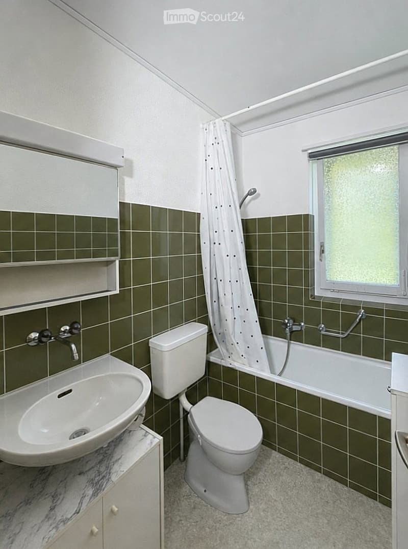 Bathroom with green tiled walls, white fixtures including a sink, toilet, and bathtub, and a window with a shower curtain.