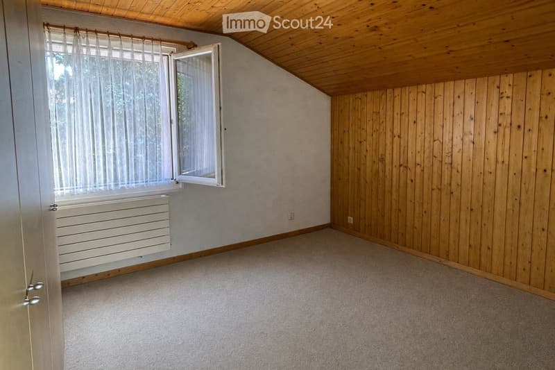 Empty room with wooden walls, carpeted floor, and a window with white curtains. The room features a white radiator below the window and a wooden ceiling.