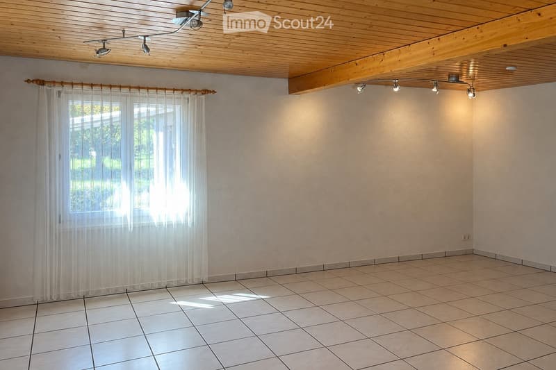Empty room with tiled floor, light-colored walls, large window with sheer curtains, and wooden ceiling with track lighting.