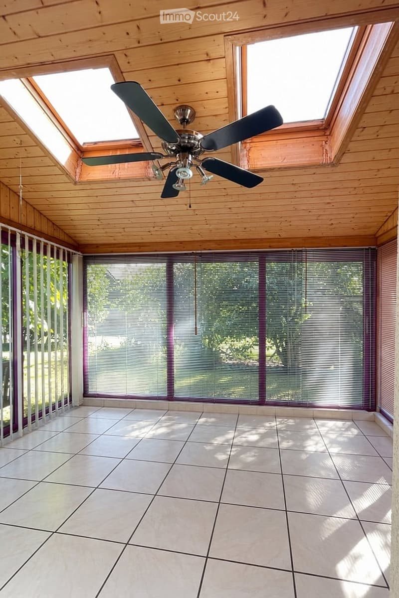 Bright, unfurnished room with wooden ceiling, large windows with blinds, and a ceiling fan. The floor is tiled, and the space is empty with no furniture.