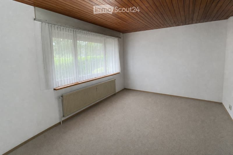 Empty room with white walls, carpeted floor, wooden baseboards, radiator, and large window with sheer curtains.