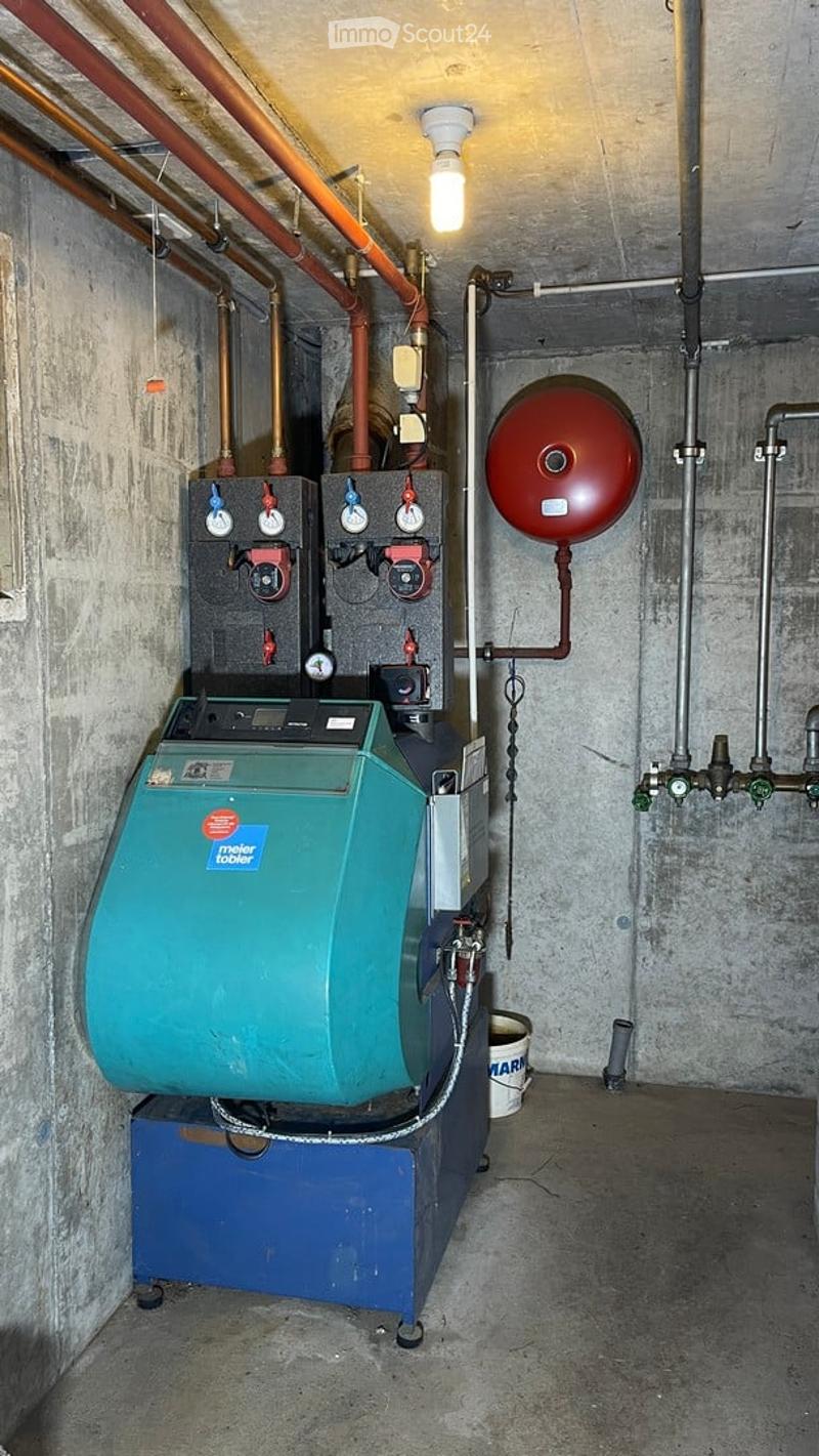 Utility room with a green boiler, pressure vessel, multiple pipes, valves, and a control panel mounted on a concrete wall.