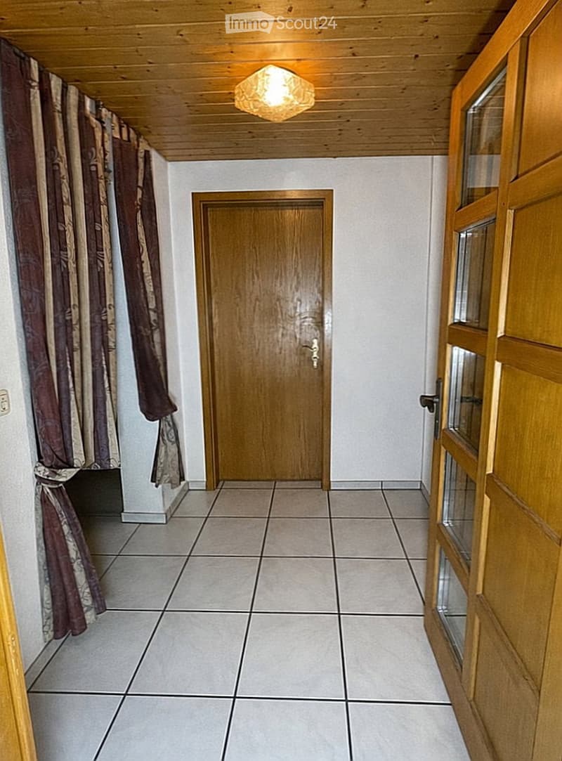 A narrow hallway with tiled flooring, wooden ceiling, and a single wooden door at the end. There is a curtain on the left side and a glass-paneled door on the right.
