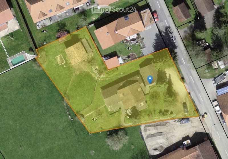 Aerial view of a large, rectangular plot of land surrounded by neighboring properties. The plot is mostly grassy with a small paved area and a small structure in the center. The surrounding area includes houses, roads, and other plots.