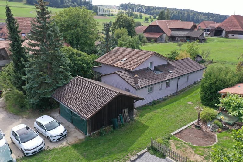 Aerial view of a two-story house with a red-tiled roof, surrounded by a well-maintained garden and several parked cars. The house is situated in a rural area with neighboring houses and open fields in the background.