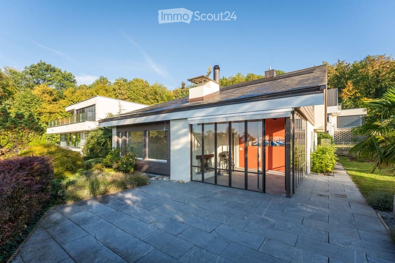Modern two-story house with a flat roof, large glass sliding doors, and a spacious patio area. The house features contemporary design with clean lines and a mix of white and dark tones.