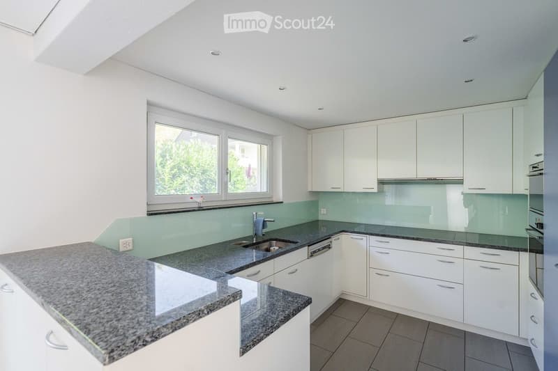Modern kitchen with white cabinets, granite countertops, a sink, and a dishwasher. Features a large window with a view of greenery.