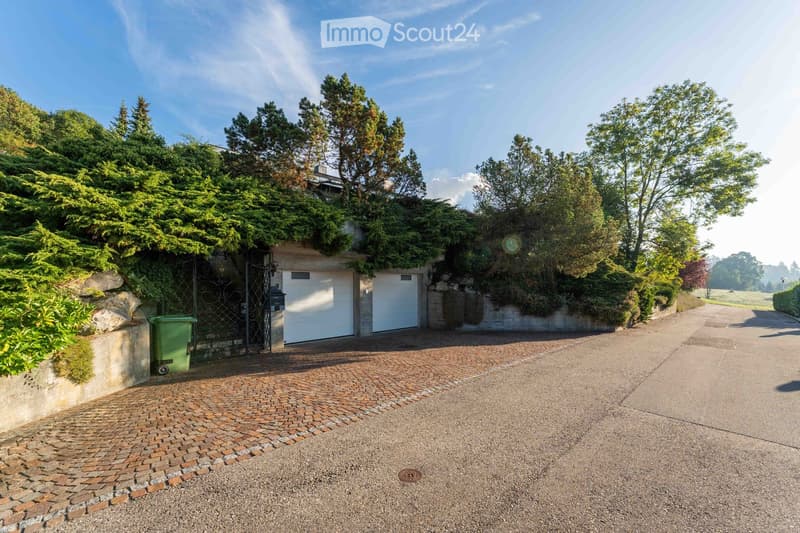 A driveway leads to a gated entrance with two garage doors, surrounded by lush greenery and trees.