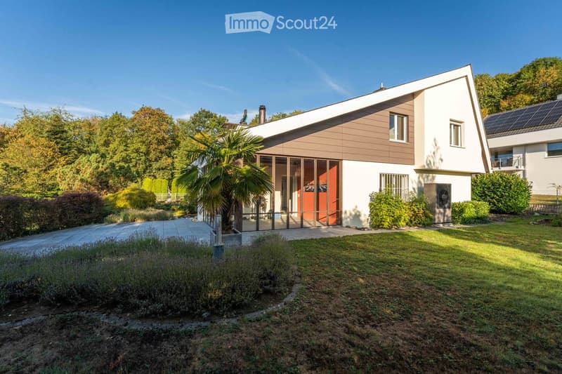 Modern two-story house with white and brown exterior, large windows, solar panels on the roof, and a well-maintained garden with a palm tree and shrubs.
