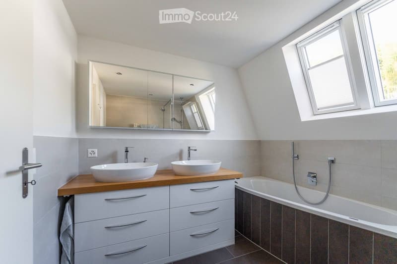 Modern bathroom with two sinks on a wooden countertop, a large mirror, a bathtub, and white tiled walls.