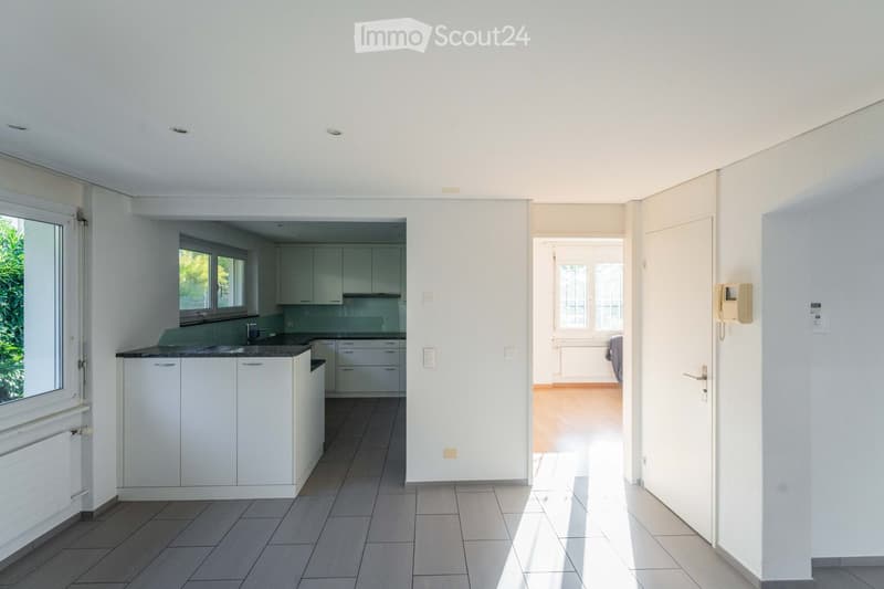 Bright, modern kitchen with white cabinets, a dark countertop, and a sink. The space is connected to a hallway with wooden flooring and white walls.
