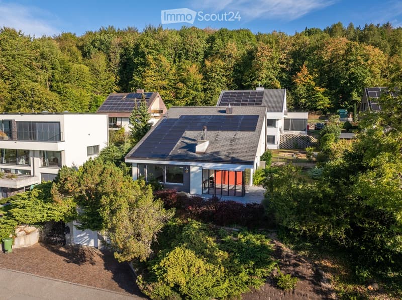 Modern two-story house with solar panels on the roof, surrounded by lush greenery and trees. Features large windows, a balcony, and a driveway.