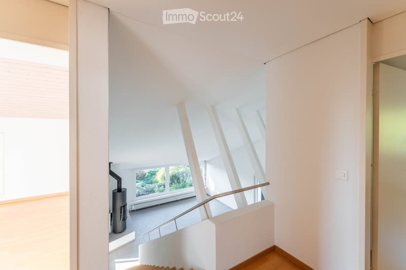Modern, minimalist hallway with white walls, wooden flooring, and a curved staircase leading upwards. Features large windows and a modern heating system on the left side.