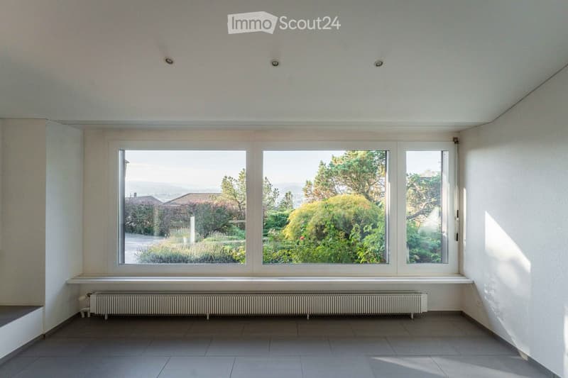 Empty room with large windows offering a view of a garden, tiled floor, radiator, and white walls.