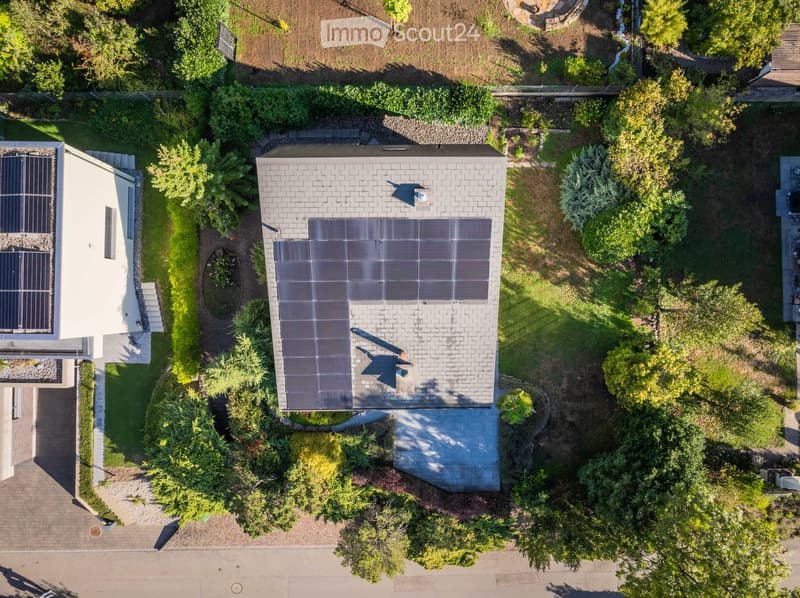 Aerial view of a two-story house with solar panels on the roof, surrounded by a well-maintained garden with trees and shrubs.