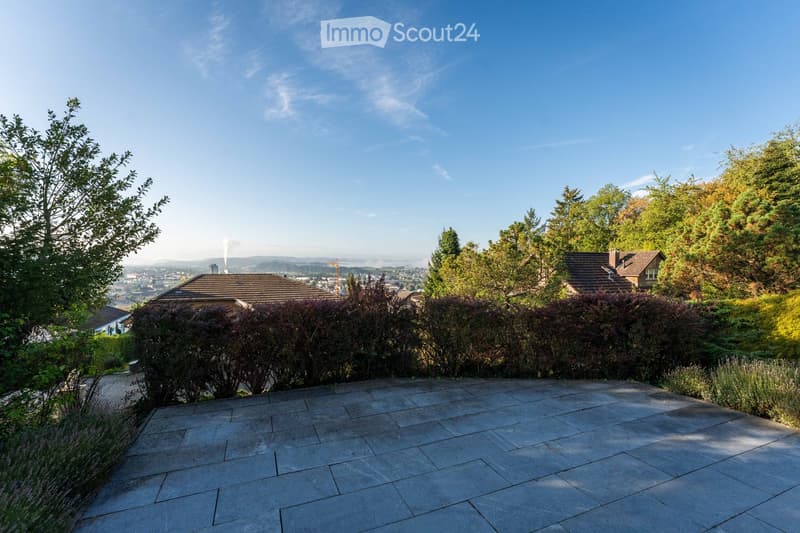 A spacious outdoor patio with a stone-tiled surface, surrounded by lush greenery and trees, offering a panoramic view of a cityscape and distant hills under a clear blue sky.