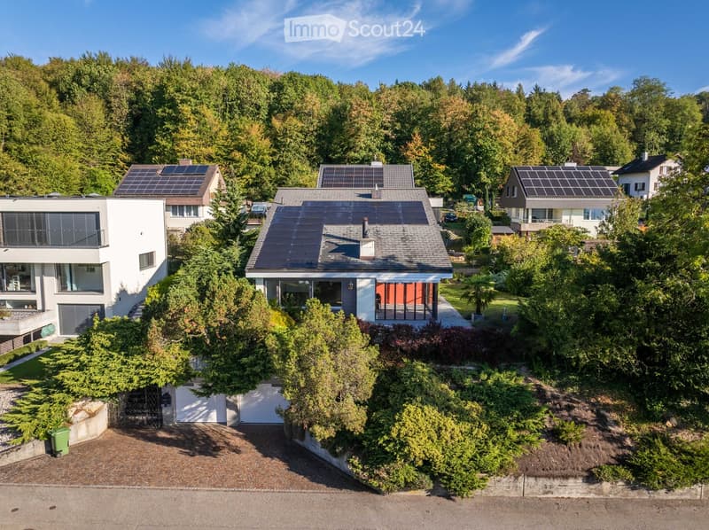 Aerial view of a modern house with solar panels on the roof, surrounded by lush greenery and trees. The house features large windows and a well-maintained garden.